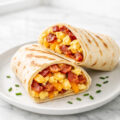 Breakfast wraps with scrambled eggs, bacon, and melted cheese cut in half on a white plate