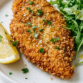 Golden brown crispy chicken Milanese cutlet on a white plate with lemon wedge and arugula