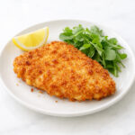 Golden brown crispy chicken Milanese cutlet on a white plate with lemon wedge and arugula