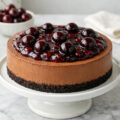 Chocolate cherry cheesecake on a white cake stand with cherry compote and whole cherries on top