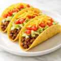 Ground beef tacos in crispy corn shells with lettuce tomato and cheese on a white plate