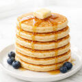Stack of fluffy homemade pancakes drizzled with maple syrup on a white plate