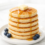 Stack of fluffy homemade pancakes drizzled with maple syrup on a white plate