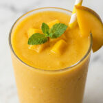 Thick creamy mango smoothie in a tall clear glass with fresh mango slice garnish on white marble surface