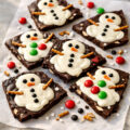 Pieces of melted snowman chocolate bark with candy eyes and pretzel arms on parchment paper