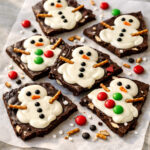 Pieces of melted snowman chocolate bark with candy eyes and pretzel arms on parchment paper